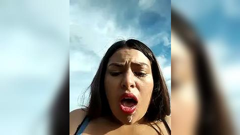 Media: Video of a young woman with dark hair, wearing a black top, with a shocked expression, drool on her face, against a cloudy sky.