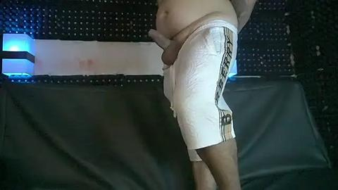 Media: Video of a light-skinned man in white shorts with a visible erection, standing in a dimly lit room with a black perforated wall and a dark gray couch.