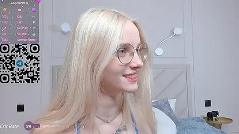 Media: Video of a smiling, fair-skinned, blonde woman with glasses and a light blue top, in a modern, light-colored bedroom with a bed, white wall, and a QR code overlay.