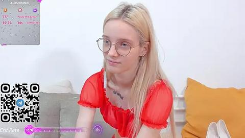 Media: Video of a fair-skinned, blonde woman with glasses and a red top, sitting on a grey couch with orange cushions in a white room.