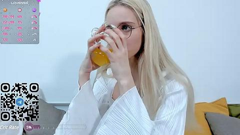 Media: Video of a blonde woman with glasses, wearing a white, long-sleeved shirt, drinking orange juice. Background features a calendar and QR code, indoor setting.