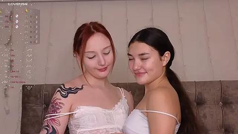 Media: Video of two young women with fair skin, one with red hair and tattoos, wearing a white lace dress, and the other with long black hair in a white tank top, smiling intimately. Background features a textured wall and a grey couch.