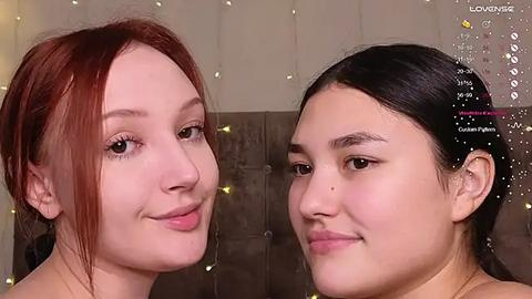 Media: Video of two young women with fair skin, one with red hair and one with black hair, smiling, against a textured wall with warm, string lights.