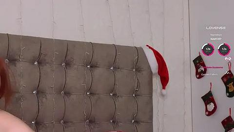 Media: Video of a gray tufted headboard with a white wall background, featuring a red Santa hat and stockings with \"LOVINA\" text, and a pink gift box.