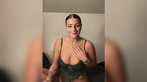 Media: Video of a fair-skinned woman with dark hair pulled back, wearing a dark green lace bodysuit, sitting on a bed, smiling.