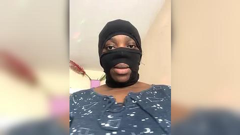 Media: Video of a woman with medium brown skin, wearing a black mask and dark patterned blouse, standing in a beige room with blurred red flowers in the background.