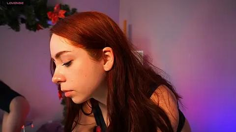 Media: A video of a young Caucasian woman with long, auburn hair and fair skin, wearing a black top, standing in a dimly lit room with Christmas decorations and purple lighting.