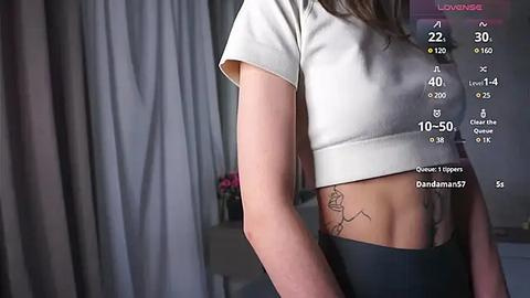 Media: Video of a slender, light-skinned woman with a short white crop top and black leggings, showcasing a tattoo on her ribcage. Background includes grey curtains and a pink flower vase.