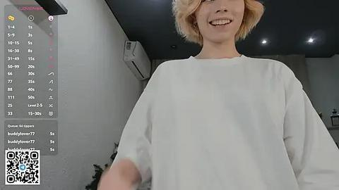 Media: Video of a smiling, light-skinned woman with short blonde hair wearing a loose white t-shirt, standing in a modern, dimly lit room with a white brick wall, an air conditioner, and a QR code in the bottom left corner.