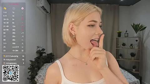 Media: A video of a blonde woman with a short bob haircut, wearing a white tank top, sticking out her tongue while covering her mouth. She's in a modern bedroom with a gray bed, green plants, and a shelf with decor.