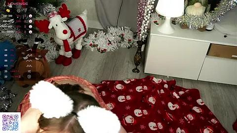 Media: A video shows a cozy living room with a white dresser, a white lamp, a red blanket with reindeer patterns, a stuffed reindeer toy, and a Christmas tree, with a person wearing a brown jacket and red mittens.