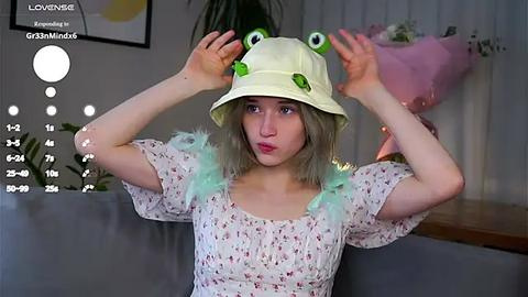 Media: Video of a young woman with light skin and shoulder-length, wavy, blonde hair, wearing a white bucket hat with green frog eyes and a pastel floral dress with feather details. She sits in a modern living room with a gray couch, pink balloons, and a potted plant in the background.