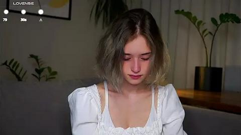 Media: Video of a young Caucasian woman with shoulder-length, light brown hair, wearing a white, off-the-shoulder blouse. She has a contemplative expression. Background features a gray couch, potted plants, and a wooden table.