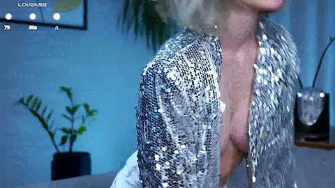 Media: A video of a blonde woman with a medium build, wearing a sequined, open-front jacket, standing in a dimly lit room with a potted plant and a blurred background.