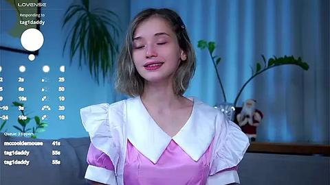 Media: A video of a young woman with fair skin, straight blonde hair, and a light pink satin dress with white ruffled collar, smiling with closed eyes, in a modern living room with potted plants and a calendar overlay.