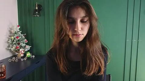 Media: Video of a young girl with long brown hair, wearing a black top, sitting indoors against a green wall, next to a decorated Christmas tree and a dark table.