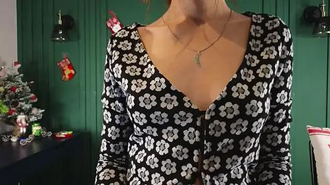 Media: Video of a woman in a black floral-patterned blouse with a deep V-neck, wearing a gold necklace, standing indoors. The background features a green wall with Christmas decorations, including a tree and ornaments.