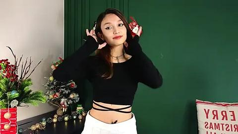 Media: Video of a young Asian woman with long brown hair, wearing a black crop top and white skirt, posing with Christmas ornaments in front of a green curtain and a decorated Christmas tree.