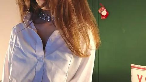 Media: Video of a woman with long, light brown hair wearing a white, button-up blouse, adorned with a floral brooch, standing in front of a green wall with a red heart decoration.