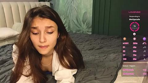 Media: Video of a young woman with long dark hair, lying on a bed, wearing a white top, in a dimly lit room with green wallpaper and a gray quilt.