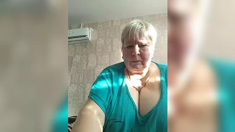 Media: A video of an older, overweight woman with short, platinum blonde hair, wearing a low-cut turquoise top, in a sunlit room with beige tiled walls and an air conditioning unit.