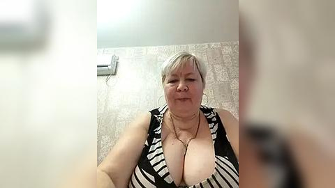 Media: A video of a middle-aged woman with short, platinum blonde hair, wearing a low-cut black and white striped dress, standing in a tiled bathroom with a beige, patterned wallpaper.