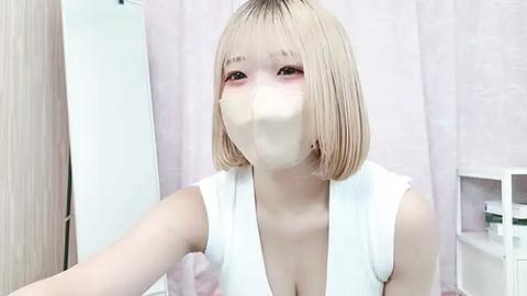 Media: Video of an East Asian woman with shoulder-length, straight blonde hair and fair skin, wearing a white sleeveless top, face mask, and blush, in a bathroom with light-colored walls and a pink curtain.