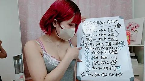 Media: Video of a young woman with red hair, wearing a face mask and gray camisole, holding a handwritten sign in Japanese about a video.