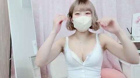 Media: Video of a young East Asian woman with short, light brown hair, wearing a white lace-trimmed camisole and a white face mask, playfully tugging her hair. The background features a pink curtain and a white vanity with a pink cushion.