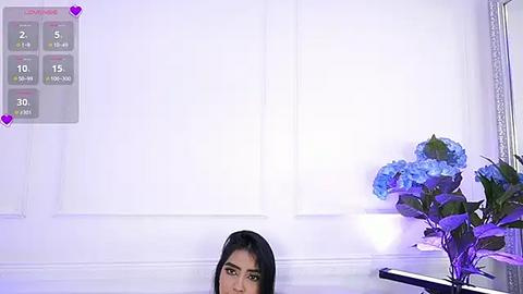 Media: Video of a woman with long black hair, partially obscured by a white wall, with a calendar and a vase of blue hydrangeas in the background.