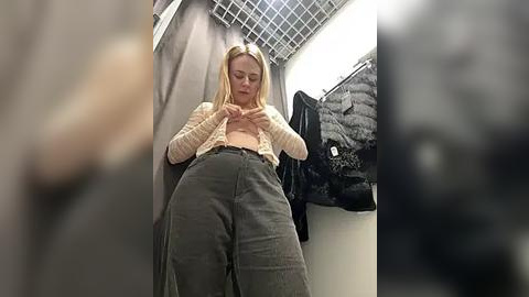 Media: Video of a young, light-skinned woman with straight blonde hair, wearing a cream knitted cardigan, unbuttoned to reveal her midriff, and high-waisted, loose-fitting, dark green pants. She stands in a small, dimly-lit changing room with metal grid ceiling, gray curtain, and hanging clothes.