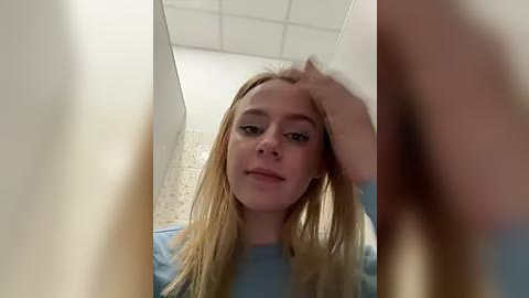 Media: A close-up video of a young Caucasian woman with long, straight blonde hair, wearing a light blue shirt, standing in a tiled bathroom. She has a neutral expression and one hand in her hair.