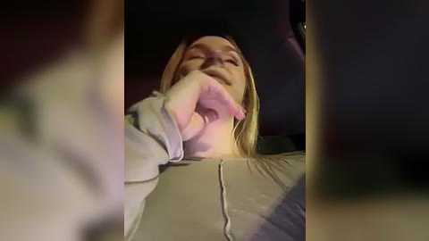 Media: Video of a blonde woman in a gray sweater, lying back in a dimly lit car, with a blurred figure in the foreground.