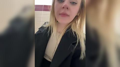 Media: A video of a young, blonde woman with fair skin, wearing a black jacket over a beige top, standing in a tiled bathroom with maroon accents. Her expression is neutral.