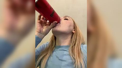 Media: Video of a young blonde woman in a blue shirt drinking from a red plastic water bottle. She appears to be in a bathroom, with blurred, indistinct figures in the background.