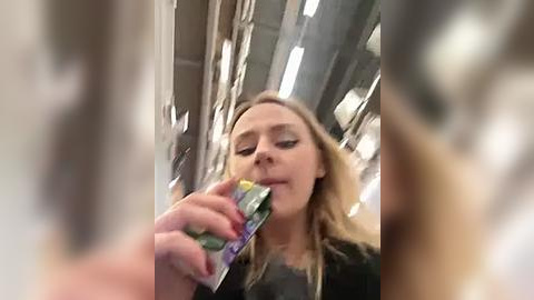 Media: A blurry video of a blonde woman eating a snack on a train, wearing a black top, with a metallic reflection visible in the background.