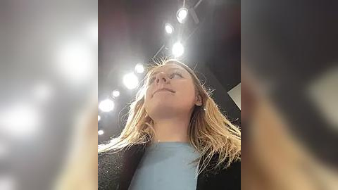 Media: A video of a blonde woman with light skin, wearing a light blue top and black jacket, looking up at bright overhead lights in a dimly lit room.