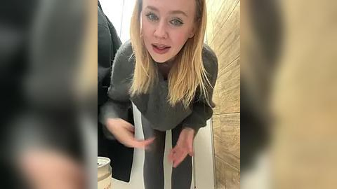 Media: Video of a young, fair-skinned blonde woman with straight hair, wearing a grey sweater and dark pants, bending over in a public restroom stall with beige tiles and a black jacket hanging nearby.