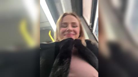 Media: Video of a smiling woman with straight blonde hair, wearing a black fur coat and pink top, inside a subway car with yellow seats and overhead lights.