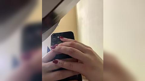 Media: A close-up video of a person's hands holding a black smartphone, partially blurred. The person has light skin and red-painted nails. The background shows a beige wall and part of a wooden table.