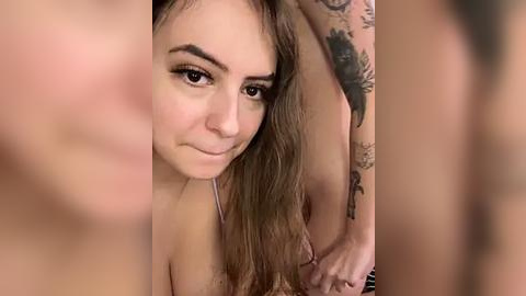 Media: Video of a young woman with fair skin, brown hair, and tattoos, topless, smiling and looking directly at the camera.