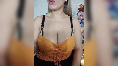 Media: Video of a blonde woman with fair skin, wearing a low-cut, orange and black spaghetti strap top, revealing ample cleavage, with a blurred background of a decorated Christmas tree.