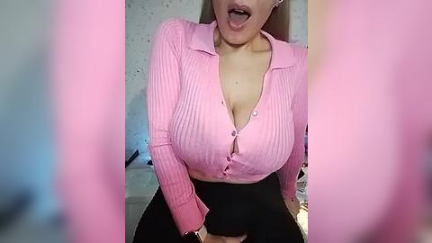 Media: Video of an Asian woman with light skin, wearing a pink ribbed button-up blouse, exposing cleavage, and black pants. She's leaning forward with her tongue out, against a blurred background.