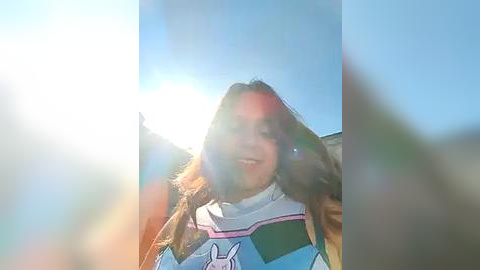 Media: A video of a smiling young woman with long brown hair, wearing a white and blue striped shirt with a bunny design, taken outdoors on a bright, sunny day, with a sun flare in the background.