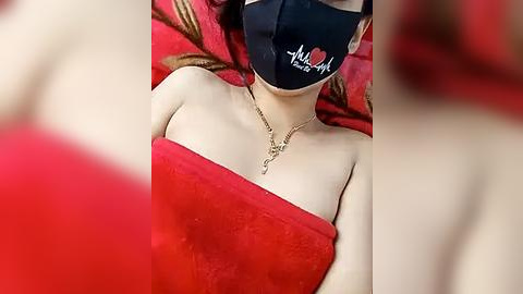 Media: Video of a fair-skinned woman with blonde hair, wearing a black face mask, red towel, and a gold necklace.