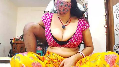 Media: Video of a South Asian woman with long black hair, wearing a pink polka-dotted top, black bra, yellow sari, and a red necklace, sitting on a bed in a cluttered room.