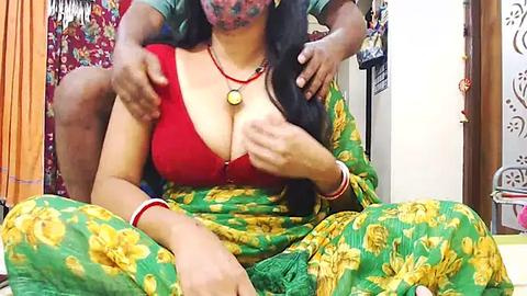 Media: Video of a South Asian woman in a green sari with yellow floral print, exposing cleavage in a red bra, sitting with two men, one behind her, in a colorful, cluttered room.