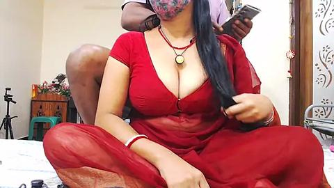 Media: A video of an Indian woman in a red sari, with large breasts, sitting on a bed, being filmed by a man behind her.