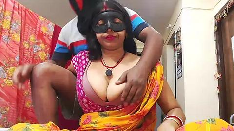 Media: Video of a dark-skinned woman with large breasts, wearing a pink and yellow saree, black blindfold, and red lipstick, being groped by a man in a striped shirt in a cluttered room.