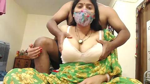 Media: A video of a dark-skinned woman with long black hair, wearing a colorful face mask, green sari, and white lace bra, sitting on a bed, revealing her breasts. The room has a wooden dresser, pink towel, and white fan in the background.